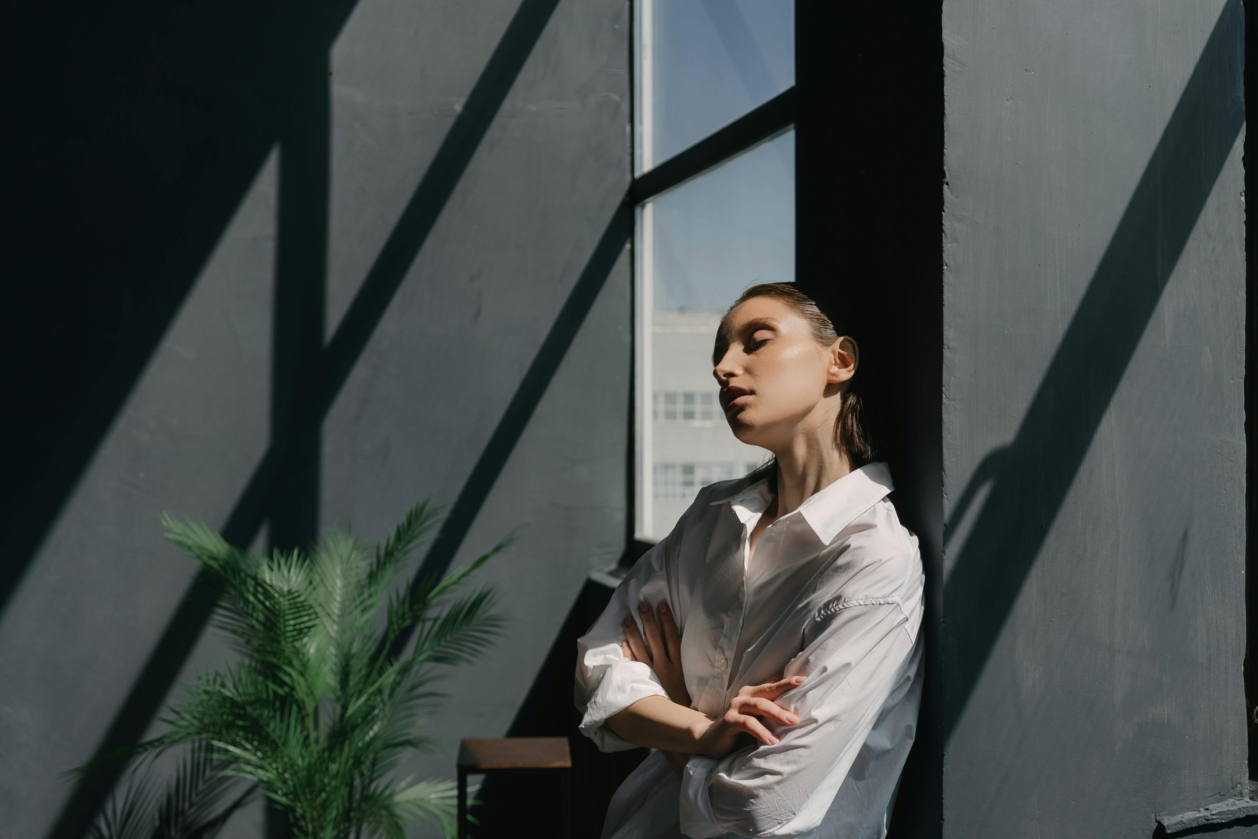 A woman in a white dress shirt leans against a black wall with closed eyes, bathed in sunlight indoors.
